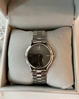 Front view of the Calvin Klein stainless steel watch with black dial, placed on a cushion inside a gray box.