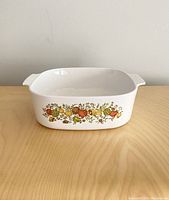 Front angled view of white CorningWare A84-B casserole dish with colorful Spice of Life vegetable and spice pattern on side, showing rectangular shape and handles