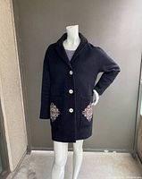 Front view of black coat showing embroidery on front pockets, three large cream buttons, and collar. Coat displayed on white mannequin with hand on hip.
