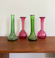 Four vintage glass vases displayed side by side on wood surface against a plain light background. Two green and two cranberry colored vases, showing their shapes and textures.