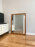 Full front view of the mid-century modern teak framed rectangular mirror leaning against a light wall on wood floor next to a white dresser and television stand.