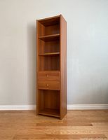 Front-side angle of tall narrow bookcase with shelves and drawers in wood grain finish.