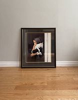 Framed print showing young person in black hood with white cat, resting against wall on floor.