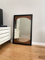 Full view of the large rectangular mirror leaning against a wall near a white media cabinet, showing the black frame and oval reflective surface with woodgrain accents top and bottom.
