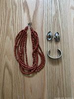 Photo of red coral beaded necklace alongside silver-tone sculptural clip-on earrings and abalone inlay cuff bracelet on wood surface.