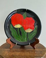 Front view of vintage USSR ceramic plate featuring embossed red tulip flowers on a black background, displayed on a wooden stand.