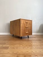 Front and side view of a mid-century wooden cabinet with three drawers and tapered legs placed on a wooden floor against a plain wall.