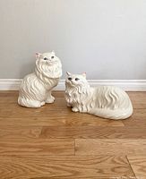Front view of two life size ceramic Persian cat statues, one sitting and one laying down, white with detailed fur texture and blue eyes.