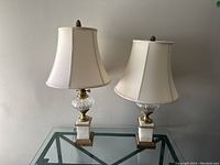 Front view of the two lamps showcasing the fabric shades, brass fittings, crystal midsections, and marble bases on a glass table.