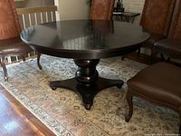 View of round black finished pedestal dining table with chairs around it, placed on patterned carpet. Top surface shows wear and scratches.