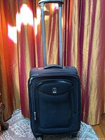 Front view of black Travelpro wheeled luggage with three zippered compartments and logo patch, standing upright with handle extended in front of curtain.