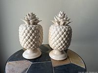 Pair of cream-colored ceramic pineapples with crackled glaze on a tiled surface, showing texture and pedestal base.