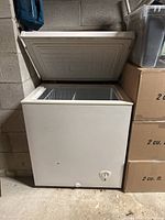 Photo showing off-white compact chest freezer with open lid, positioned next to boxes, on a concrete floor.