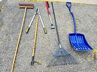 Photo showing a wooden-handled push broom, hedge shears with yellow and black handles, cultivator, leaf rake, and blue snow shovel laid on pavement.