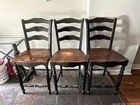 Three ladder back bar stools showing caramel wooden seats, black painted carved legs and ladder style backs with floral painted top rail.