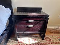 Photo of one dark wood nightstand with two drawers and metal handles showing surface wear and fading paint on top.