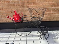 Photo of black metal Penny Farthing bicycle planter with red and white polka dot metal watering can in front basket, showing full bike profile.