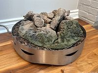Oval stainless steel gas outdoor fireplace base with ventilation cutouts, filled with faux ceramic logs resembling natural wood with ash and moss textures. Attached flexible metal gas hose visible.