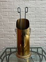 Front view of polished brass fireplace tool holder with two black metal firewood tongs inside.