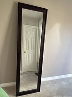 Tall standing mirror leaning against a beige wall, dark wood frame visible.