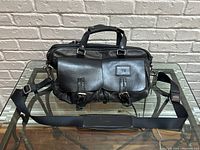 Front view of black leather Tumi travel bag on glass table beneath white brick wall.