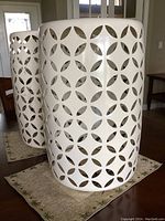 Two white metal cylindrical plant stands with geometric cut-outs shown on floor mat inside a home.