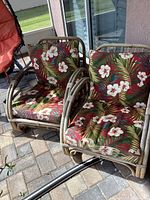 Pair of bamboo papasan chairs with Hawaiian floral cushions placed outdoors on a stone patio. Chairs display rounded bamboo framing and thick colorful cushions with hibiscus-style flowers and palm leaves.