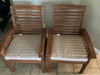 Two Bermuda style wooden chairs with beige cushions photographed from the front showing slatted backs and armrests on tiled flooring.