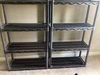 Two black plastic shelving units, each with four shelves, standing side by side on a carpeted floor against a light-colored wall.