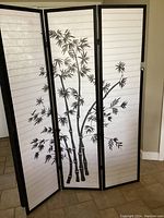 The photo shows a three-panel black framed Oriental Shoji screen standing upright. Each panel displays a white translucent section with horizontal ribbing and a black bamboo design spanning across the panels.