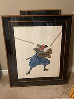 Framed samurai print artwork in koa wood frame shown upright against wall, featuring detailed samurai figure in traditional attire and a black mat with red accent.