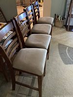 Four wooden bar stools lined up on carpet, showing ladder-back design and worn beige cushioned seats.