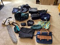 Photo shows 15 assorted bags in different styles and colors laid on carpeted floor including floral tote, navy/tan satchel, green duffel, and smaller pouches.