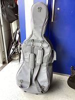 Front view of gray cello soft case showing backpack-style padded straps and attachment rings on gray fabric material.