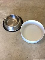 Photo showing both the stainless steel dog bowl with black rubber base and the white ceramic dog bowl on a concrete surface.
