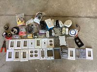 Box with various electrical parts including white wall plates, electrical outlets, breakers, wire caps, tape, timer switch, metal box, and small tool