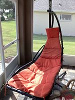 View of black metal frame free standing hammock lounge with an orange padded cushion, positioned near a screened porch with outdoor scenery.