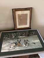 Wooden frame leaning against the wall with a framed art picture of two German Shepherd dogs lying outdoors. Photo shows the artwork, frame style, and matting.