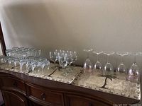Wide shot of all 32 pieces showing 20 matching drinking glasses, 7 glass wine glasses, and 5 decorated plastic wine glasses arranged on wooden cabinet.