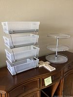 Photo showing a 4-tier white plastic office basket organizer, a 3-tier round glass and metal lazy Susan, and a light-colored decorative cross on a wooden furniture surface against a beige wall.
