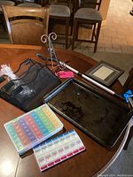 Photo showing full lot including black mesh office organizer, colorful and clear plastic weekly pill boxes, metal grabber tool with blue grips, pink luggage tag, small metal frame easel, wooden dog picture frame, and flat metal cooking sheet pan with dark stains.