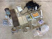 Overview of assorted garage hardware including steel base plates, pulleys, chains, washers, screws, hooks, carabiners, and other fastening components laid on concrete floor.