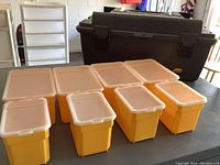 Eight yellow plastic storage bins with white lids arranged in front of a large black Plano plastic toolbox.