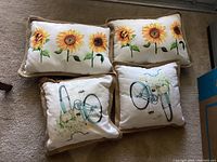 Four decorative pillows on carpet: two sunflower design rectangular pillows and two bicycle design square pillows with beige fringe edging.