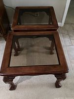 Pair of square wooden end tables with inset glass tops and carved shell-themed claw feet, medium brown finish, standing side by side on carpeted floor near tiled floor.