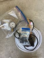Overview photo showing the submersible pump with attached power cord, white coiled hose, pressure gauge in bag, and mounting hardware in small packets.