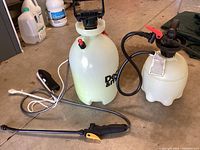 Photo showing two weed sprayers including a larger 2 gallon Decks & More sprayer with spray wand and pump handle, and a smaller 1 gallon Gilmore sprayer with pump handle and flexible hose