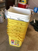Six yellow 35 lb capacity plastic buckets stacked with white lids on top, located in a garage area with other household containers visible in the background.