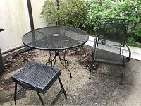 Full view of round table, chair, and small side table/stool on outdoor concrete surface with greenery in background.