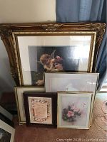 Various framed art including a large mother and child print in an ornate gold frame, smaller floral and text prints in wooden and gold frames, arranged leaning against a wall.